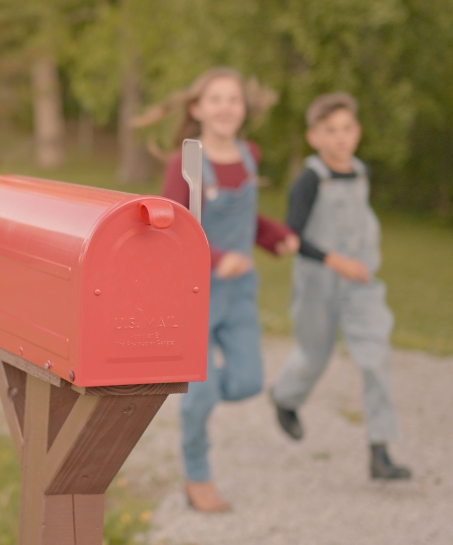 Two children in overalls running toward a red mailbox on a rural path